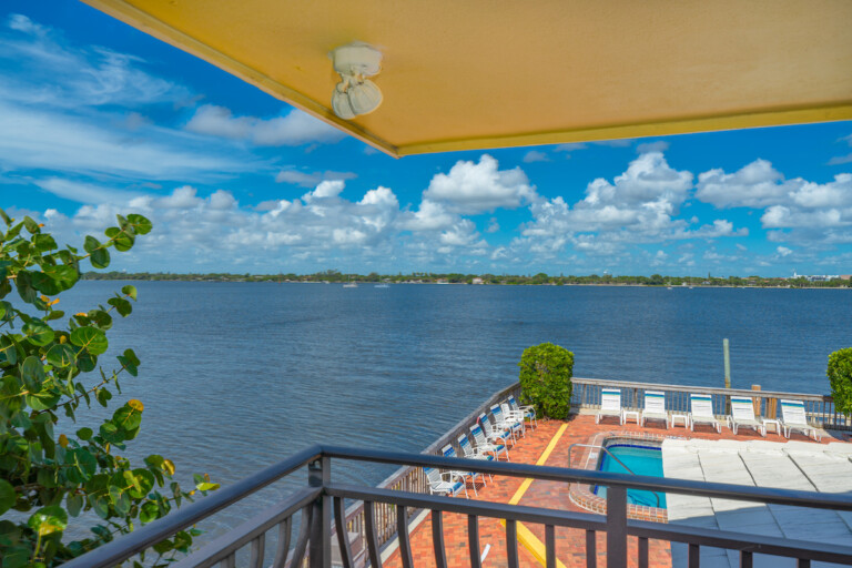 View from a covered private balcony facing down at a waterfront community pool and brick patio, with an expansive Intracoastal and bright blue sky in the background.