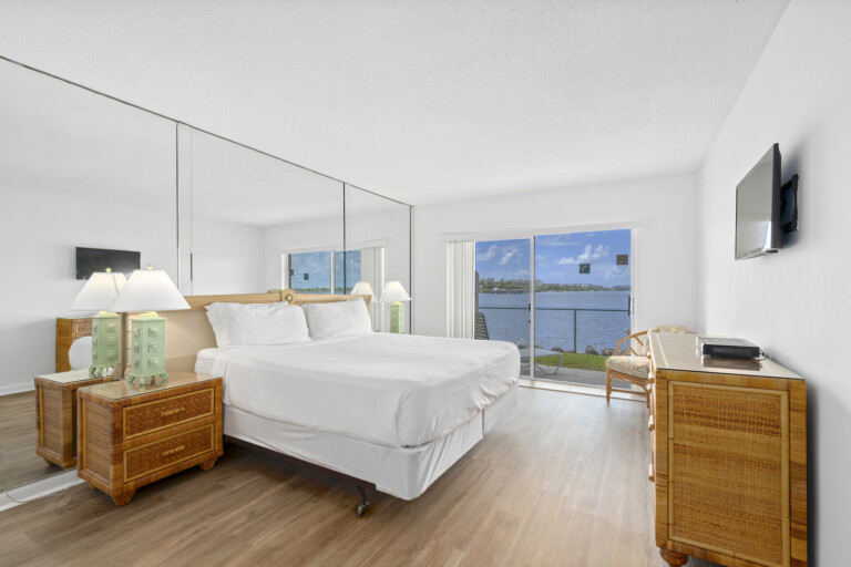 Waterfront bedroom with a large mirrored wall, rattan dresser and nightstands, and a sliding glass door.