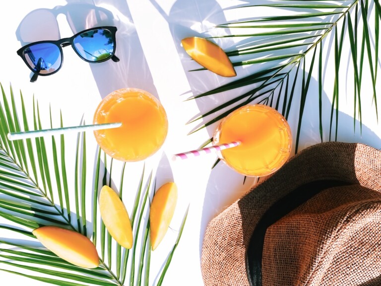 Summer flat lay of two glasses of fresh mango juice with striped straws, mango slices, sunglasses, a straw hat, and palm leaves on a bright white surface.