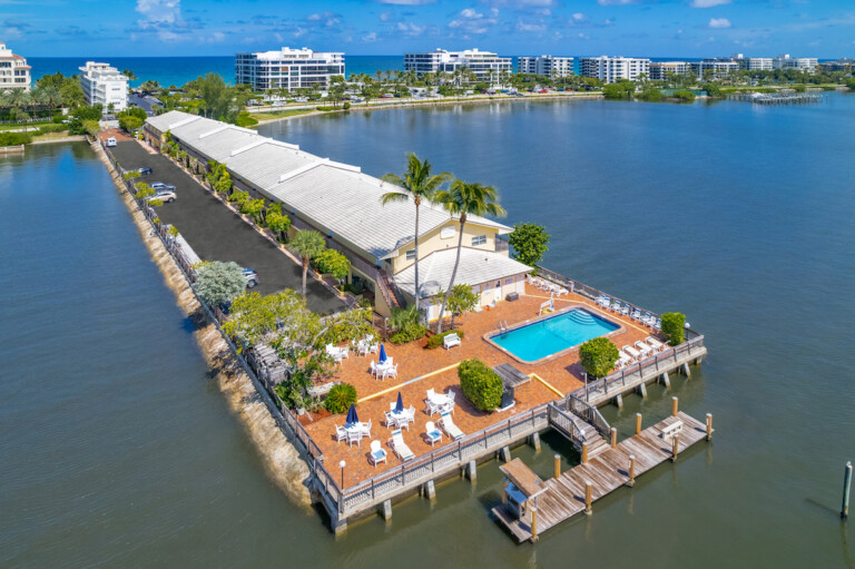 Aerial panoramic of a coastal resort on a narrow peninsula, featuring a swimming pool, brick patio, and wooden boat dock, with luxury condos and the Atlantic Ocean in the background.