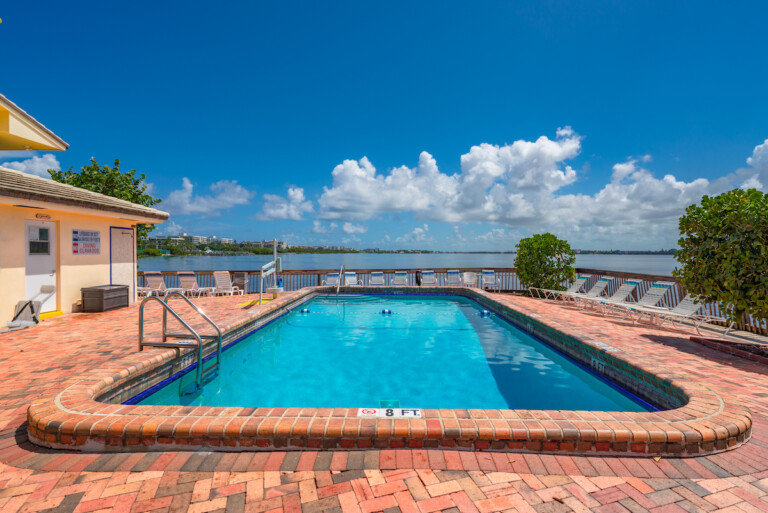 Waterfront community pool on a brick patio, surrounded by lounge chairs and overlooking the Intracoastal Waterway under a bright blue sky.