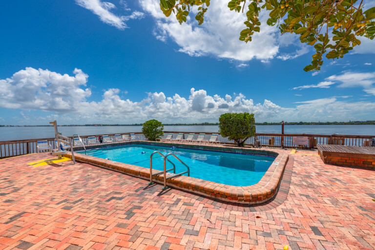 Waterfront swimming pool on a brick paver patio with lounge chairs, overlooking a wide river under a bright blue sky with white clouds.