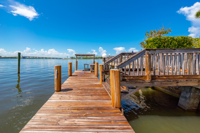 Wooden boat dock and pier extending into a the intracoastal with a bridge in the distance under a bright blue sky.