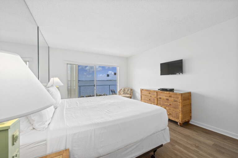 Bright waterfront bedroom with white bedding, a rattan dresser, a wall-mounted TV, and sliding glass doors facing the intracoastal.